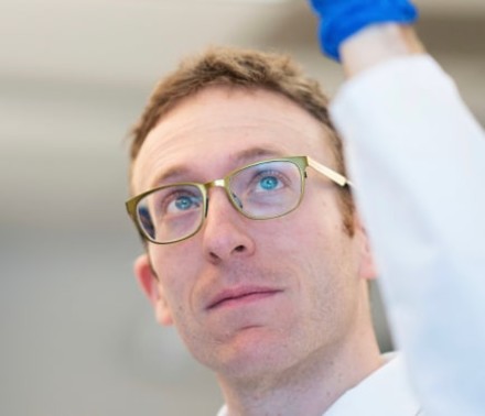 Close-up lifestyle portrait of a male scientist in a white lab coat and glasses holding up and inspecting liquid in a vial.