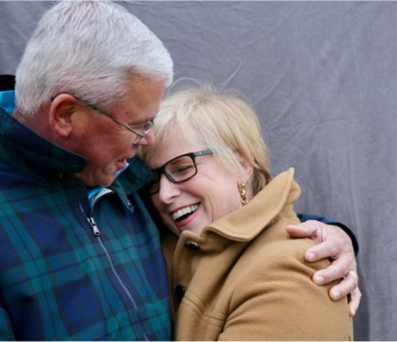 Natural portrait of a middle-aged cancer patient laughing and being embraced by her husband in front of a grey backdrop.