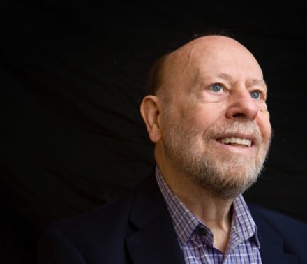 Naturally lit portrait of a senior male cancer patient smiling thoughtfully in front of a black backdrop.