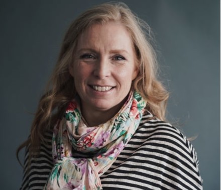 Naturally lit portrait of a middle-aged female biotech executive wearing a flowery scarf and smiling in front of a dark bluish-gray background.