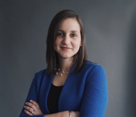 Portrait of a young female staff member in a blue blazer smiling as she looks at through the lens of the camera.