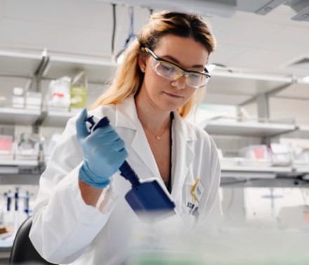 Lifestyle portrait of a middle-aged female scientist smiling for the camera in a blue lab coat, hair net, and protective glasses.