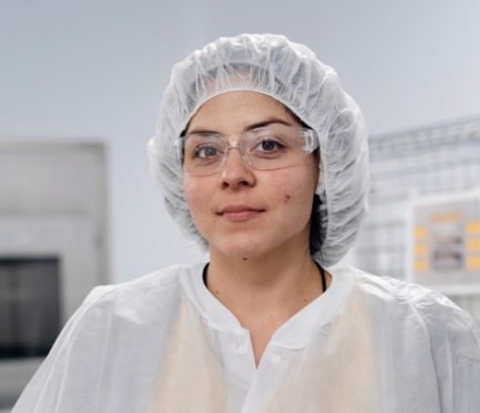 Lifestyle portrait of a young female scientist looking at the camera and smiling slightly in protective clothing, hair net, and protective glasses.