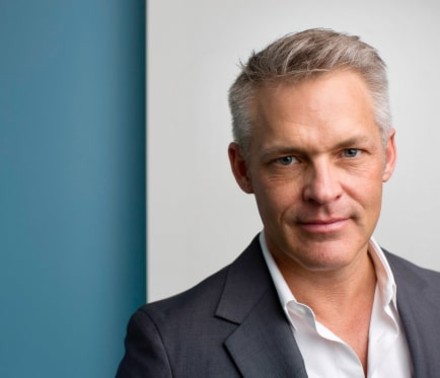 Naturally lit portrait of a male biotech executive in front of a blue and white background confidently looking into the camera lens.